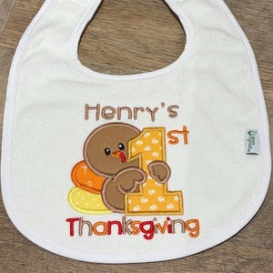 Puede incluir: Un babero blanco con un dise&ntilde;o bordado de un pavo marr&oacute;n sosteniendo un n&uacute;mero uno grande de color amarillo. El texto Henry's 1st Thanksgiving est&aacute; bordado en hilo marr&oacute;n y rojo. Una peque&ntilde;a etiqueta verde en el lateral muestra la marca Green Sprouts.