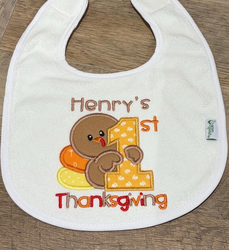 Puede incluir: Un babero blanco con un dise&ntilde;o bordado de un pavo marr&oacute;n sosteniendo un n&uacute;mero uno grande de color amarillo. El texto Henry's 1st Thanksgiving est&aacute; bordado en hilo marr&oacute;n y rojo. Una peque&ntilde;a etiqueta verde en el lateral muestra la marca Green Sprouts.