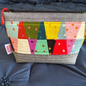 Pu&ograve; includere: Un astuccio con cerniera fatto a mano e colorato con un design patchwork. L'astuccio presenta una variet&agrave; di tessuti nei toni del rosso, giallo, verde acqua e rosa, con dettagli a stella. La parte superiore e inferiore sono grigie e il tirazip &egrave; rosso.