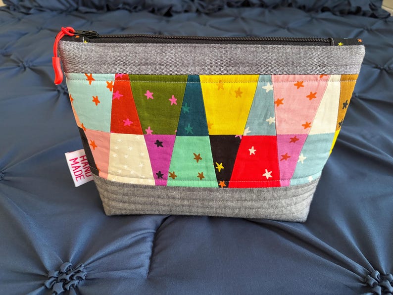 Pu&ograve; includere: Un astuccio con cerniera fatto a mano e colorato con un design patchwork. L'astuccio presenta una variet&agrave; di tessuti nei toni del rosso, giallo, verde acqua e rosa, con dettagli a stella. La parte superiore e inferiore sono grigie e il tirazip &egrave; rosso.