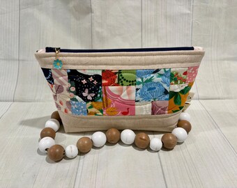 May include: A quilted fabric pouch featuring a colorful patchwork pattern of floral and butterfly motifs. The pouch has a beige top and bottom border, a dark blue zipper, and a small blue flower charm. A beaded handle with alternating white and brown wooden beads is attached.