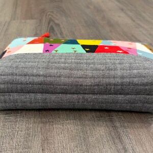 Pu&ograve; includere: Un astuccio rettangolare grigio trapuntato con una parte superiore patchwork colorata. Il patchwork presenta triangoli e stelle in vari colori, tra cui rosa, giallo e turchese. Una piccola etichetta rossa con testo bianco &egrave; attaccata al lato.