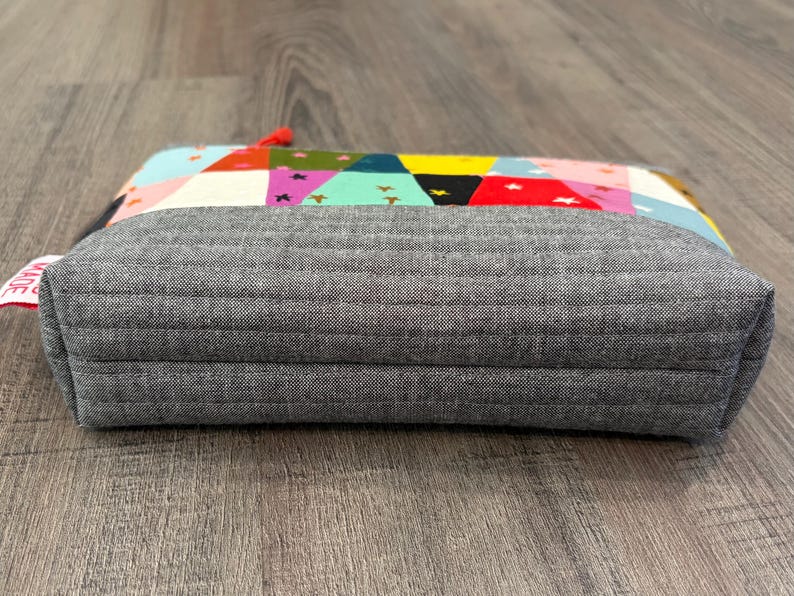 Pu&ograve; includere: Un astuccio rettangolare grigio trapuntato con una parte superiore patchwork colorata. Il patchwork presenta triangoli e stelle in vari colori, tra cui rosa, giallo e turchese. Una piccola etichetta rossa con testo bianco &egrave; attaccata al lato.