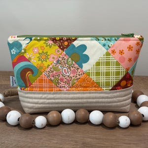 K&ouml;nnte beinhalten: Eine gesteppte Rei&szlig;verschlusstasche mit einem Patchwork-Design aus bunten Blumen-, Paisley- und Karostoffen in Gelb, Orange, Blau und Gr&uuml;n. Die Basis besteht aus beigem Stoff mit horizontalen N&auml;hten, akzentuiert durch eine Kette aus Holzperlen.
