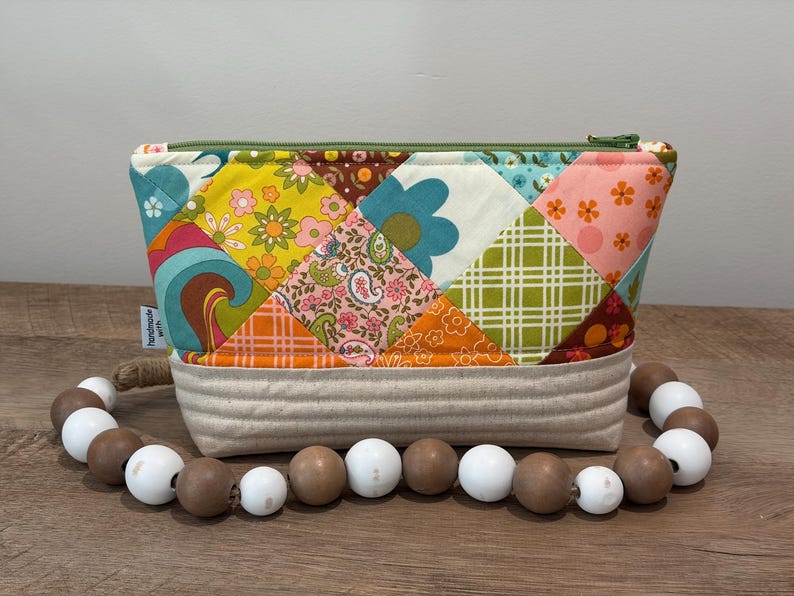 K&ouml;nnte beinhalten: Eine gesteppte Rei&szlig;verschlusstasche mit einem Patchwork-Design aus bunten Blumen-, Paisley- und Karostoffen in Gelb, Orange, Blau und Gr&uuml;n. Die Basis besteht aus beigem Stoff mit horizontalen N&auml;hten, akzentuiert durch eine Kette aus Holzperlen.