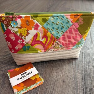 K&ouml;nnte beinhalten: Eine Rei&szlig;verschlusstasche mit einem Patchwork-Design aus floralen und geometrischen Stoffen in Pink, Orange, Gr&uuml;n und Blau, mit einer beigen, gesteppten Basis und einem Blumenanh&auml;nger am Rei&szlig;verschluss. Ein kleiner Stapel Stoffquadrate mit der Aufschrift Happy Daze liegt daneben.