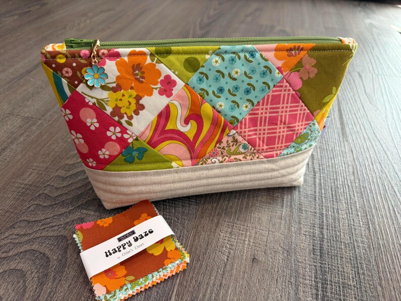 K&ouml;nnte beinhalten: Eine Rei&szlig;verschlusstasche mit einem Patchwork-Design aus floralen und geometrischen Stoffen in Pink, Orange, Gr&uuml;n und Blau, mit einer beigen, gesteppten Basis und einem Blumenanh&auml;nger am Rei&szlig;verschluss. Ein kleiner Stapel Stoffquadrate mit der Aufschrift Happy Daze liegt daneben.