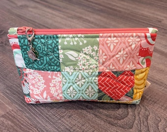 Puede incluir: Un estuche acolchado rectangular con cremallera que presenta un diseño de patchwork con varios patrones florales y geométricos en tonos rosa, verde, azul cerceta, rojo y amarillo. Un dije de metal plateado en forma de trébol cuelga del tirador.