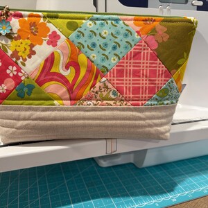 K&ouml;nnte beinhalten: Ein gestepptes Rei&szlig;verschlusst&auml;schchen mit einem Patchwork-Design aus verschiedenen Blumen-, Karomustern und geometrischen Stoffen in Orange, Pink, Gr&uuml;n und Blau. Der Boden besteht aus beigem Stoff mit horizontalen N&auml;hten, und am gr&uuml;nen Rei&szlig;verschluss befindet sich ein kleiner blauer Blumenanh&auml;nger.
