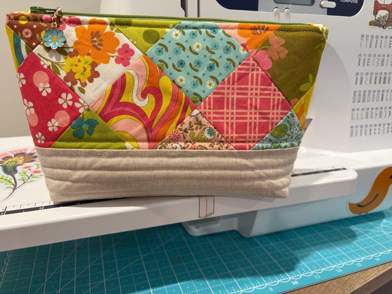 K&ouml;nnte beinhalten: Ein gestepptes Rei&szlig;verschlusst&auml;schchen mit einem Patchwork-Design aus verschiedenen Blumen-, Karomustern und geometrischen Stoffen in Orange, Pink, Gr&uuml;n und Blau. Der Boden besteht aus beigem Stoff mit horizontalen N&auml;hten, und am gr&uuml;nen Rei&szlig;verschluss befindet sich ein kleiner blauer Blumenanh&auml;nger.