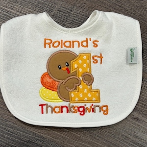 Puede incluir: Un babero blanco para beb&eacute; con un pavo marr&oacute;n bordado que sostiene un n&uacute;mero uno grande de color amarillo. El texto dice Roland's 1st Thanksgiving en hilo naranja y rojo. Una peque&ntilde;a etiqueta verde en el lateral muestra la marca Green Sprouts.