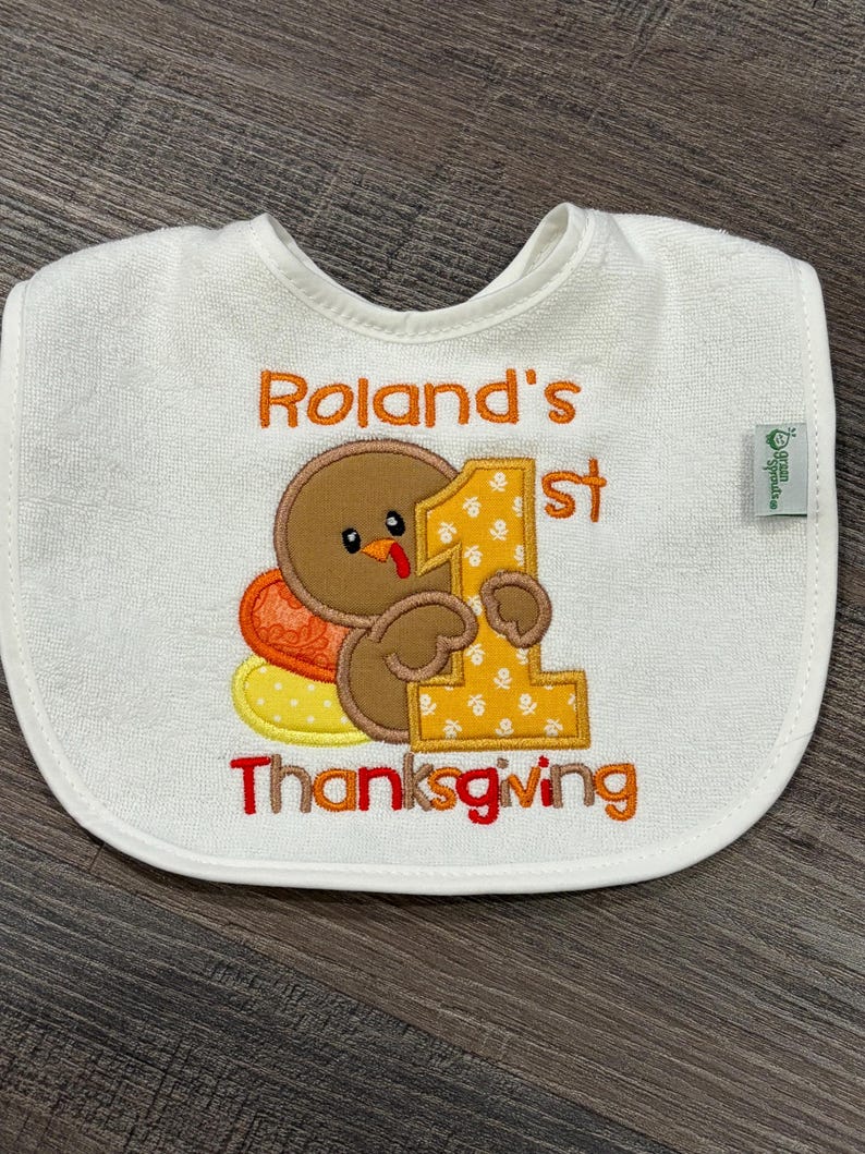 Puede incluir: Un babero blanco para beb&eacute; con un pavo marr&oacute;n bordado que sostiene un n&uacute;mero uno grande de color amarillo. El texto dice Roland's 1st Thanksgiving en hilo naranja y rojo. Una peque&ntilde;a etiqueta verde en el lateral muestra la marca Green Sprouts.
