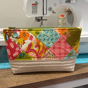 K&ouml;nnte beinhalten: Eine Rei&szlig;verschlusstasche mit einem bunten Patchwork-Design aus verschiedenen Blumen-, geometrischen und Karomustern in Gr&uuml;n-, Orange-, Rosa- und Blaut&ouml;nen. Der untere Teil besteht aus beigem Stoff mit horizontalen Steppn&auml;hten.