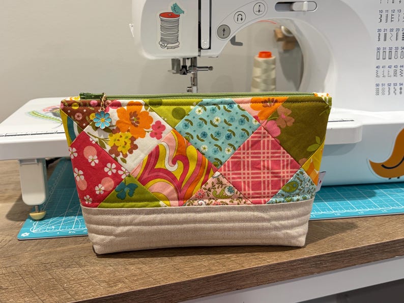 K&ouml;nnte beinhalten: Eine Rei&szlig;verschlusstasche mit einem bunten Patchwork-Design aus verschiedenen Blumen-, geometrischen und Karomustern in Gr&uuml;n-, Orange-, Rosa- und Blaut&ouml;nen. Der untere Teil besteht aus beigem Stoff mit horizontalen Steppn&auml;hten.