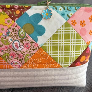 K&ouml;nnte beinhalten: Eine Rei&szlig;verschlusstasche mit einem Patchwork-Design aus verschiedenen Blumen-, Paisley- und Karostoffquadraten in den Farben Rosa, Orange, Gr&uuml;n, Blau und Braun. Die Oberkante hat einen gr&uuml;nen Rei&szlig;verschluss mit einem hellblauen Blumenanh&auml;nger und einen beigen Boden.