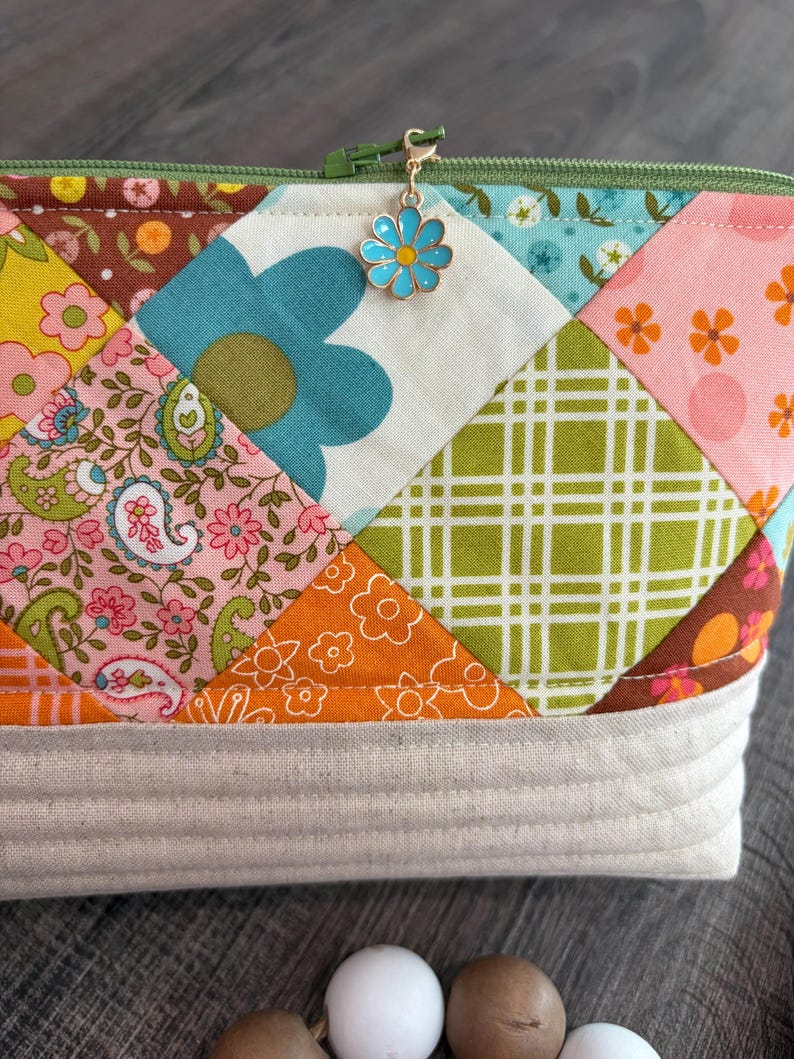 K&ouml;nnte beinhalten: Eine Rei&szlig;verschlusstasche mit einem Patchwork-Design aus verschiedenen Blumen-, Paisley- und Karostoffquadraten in den Farben Rosa, Orange, Gr&uuml;n, Blau und Braun. Die Oberkante hat einen gr&uuml;nen Rei&szlig;verschluss mit einem hellblauen Blumenanh&auml;nger und einen beigen Boden.