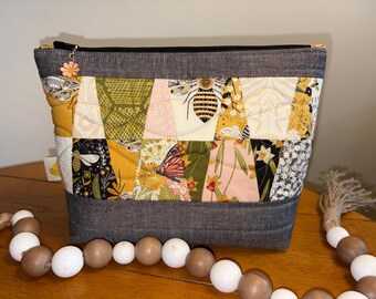 May include: A zippered pouch featuring a patchwork design of mustard yellow, olive green, black, and cream fabrics with bee and butterfly motifs. The top and bottom bands are dark gray fabric. A small orange flower charm is attached to the zipper pull.