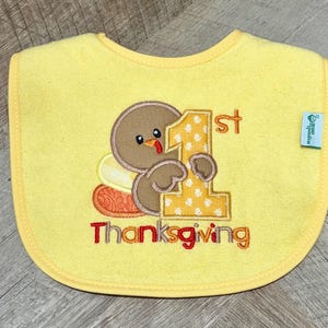 Puede incluir: Un babero amarillo para beb&eacute; con un pavo marr&oacute;n bordado que sostiene un n&uacute;mero uno grande de color amarillo con patrones blancos. El texto 1st Thanksgiving est&aacute; bordado debajo en rojo y naranja. Una peque&ntilde;a etiqueta verde est&aacute; en el lado derecho.