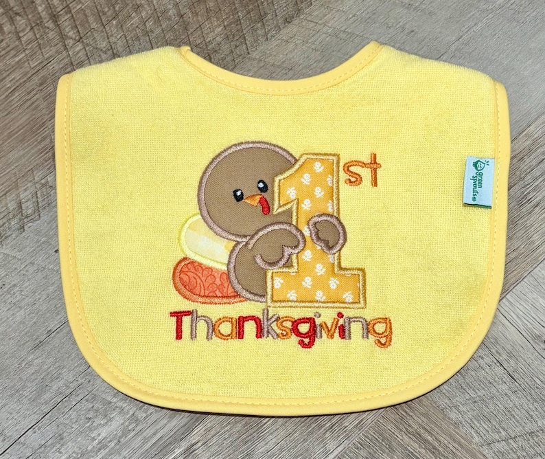 Puede incluir: Un babero amarillo para beb&eacute; con un pavo marr&oacute;n bordado que sostiene un n&uacute;mero uno grande de color amarillo con patrones blancos. El texto 1st Thanksgiving est&aacute; bordado debajo en rojo y naranja. Una peque&ntilde;a etiqueta verde est&aacute; en el lado derecho.