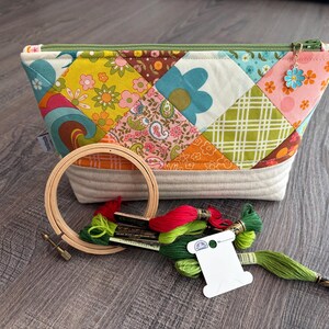 K&ouml;nnte beinhalten: Ein gestepptes Rei&szlig;verschlusst&auml;schchen mit einem bunten Patchwork-Muster aus Blumen- und geometrischen Drucken, versehen mit einem gr&uuml;nen Rei&szlig;verschluss und einem kleinen blauen Blumenanh&auml;nger. Daneben liegen ein Stickrahmen aus Holz und Stickgarn.