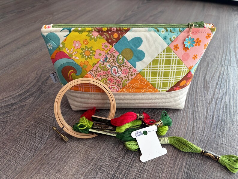 K&ouml;nnte beinhalten: Ein gestepptes Rei&szlig;verschlusst&auml;schchen mit einem bunten Patchwork-Muster aus Blumen- und geometrischen Drucken, versehen mit einem gr&uuml;nen Rei&szlig;verschluss und einem kleinen blauen Blumenanh&auml;nger. Daneben liegen ein Stickrahmen aus Holz und Stickgarn.