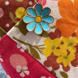 K&ouml;nnte beinhalten: Ein Stoffbeutel mit einem Patchwork-Design aus Blumenmustern in den Farben Pink, Orange, Gelb und Braun. Ein goldfarbener Metallanh&auml;nger in Form eines G&auml;nsebl&uuml;mchens mit hellblauen Bl&uuml;tenbl&auml;ttern und gelber Mitte ist am Rei&szlig;verschluss befestigt.
