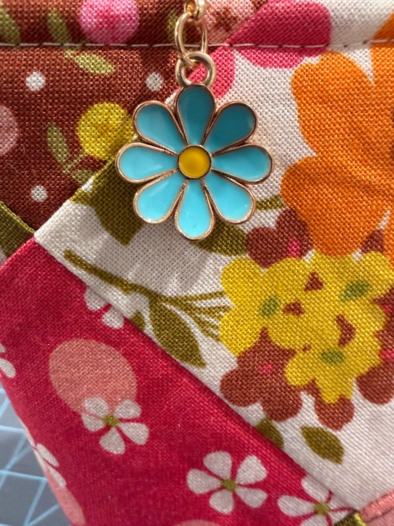 K&ouml;nnte beinhalten: Ein Stoffbeutel mit einem Patchwork-Design aus Blumenmustern in den Farben Pink, Orange, Gelb und Braun. Ein goldfarbener Metallanh&auml;nger in Form eines G&auml;nsebl&uuml;mchens mit hellblauen Bl&uuml;tenbl&auml;ttern und gelber Mitte ist am Rei&szlig;verschluss befestigt.