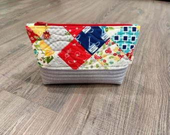 May include: A quilted zippered pouch featuring a patchwork top section with various colorful fabric patterns including red, blue, yellow, and white, and a solid gray bottom section. A small red flower charm is attached to the red zipper pull.