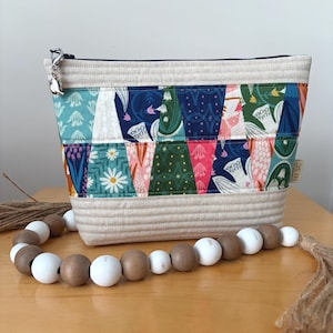 May include: A quilted zipper pouch featuring a colorful patchwork pattern of floral and geometric prints in shades of teal, navy, green, and pink, with cream-colored fabric panels at the top and bottom and a silver metal charm on the zipper pull.