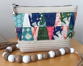 May include: A quilted zipper pouch featuring a colorful patchwork pattern of floral and geometric prints in shades of teal, navy, green, and pink, with cream-colored fabric panels at the top and bottom and a silver metal charm on the zipper pull.