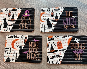 May include: Four rectangular quilted fabric mats featuring Halloween motifs like black cats, witch hats, and potions. Each mat is split diagonally between a white patterned side and a solid black side with embroidered text: SIT A SPELL, THE WITCH IS IN, MORE BOOS PLEASE, and GHOULS NIGHT OUT.