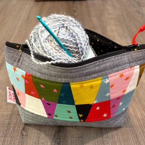 Pu&ograve; includere: Una borsa per filati fatta a mano in tessuto grigio, con un colorato design patchwork con stelle. La borsa contiene un gomitolo di filato azzurro e grigio e un uncinetto turchese. &Egrave; visibile un tirazip rosso.