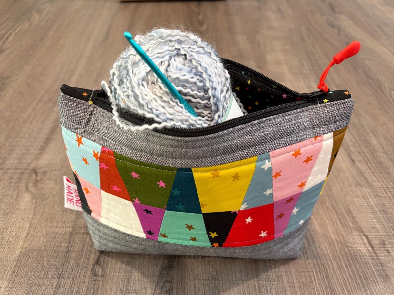 Pu&ograve; includere: Una borsa per filati fatta a mano in tessuto grigio, con un colorato design patchwork con stelle. La borsa contiene un gomitolo di filato azzurro e grigio e un uncinetto turchese. &Egrave; visibile un tirazip rosso.