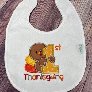 Puede incluir: Un babero blanco con el bordado de un pavo marr&oacute;n que sostiene un n&uacute;mero uno grande de color amarillo. El texto 1st Thanksgiving est&aacute; bordado en naranja y rojo. Una peque&ntilde;a etiqueta verde en el lateral muestra el texto green sprouts.