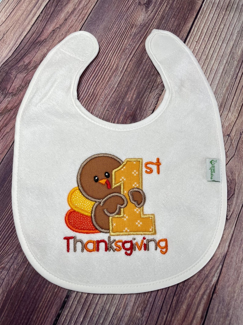 Puede incluir: Un babero blanco con el bordado de un pavo marr&oacute;n que sostiene un n&uacute;mero uno grande de color amarillo. El texto 1st Thanksgiving est&aacute; bordado en naranja y rojo. Una peque&ntilde;a etiqueta verde en el lateral muestra el texto green sprouts.