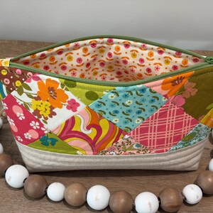 K&ouml;nnte beinhalten: Ein Rei&szlig;verschlusst&auml;schchen mit einer Patchwork-Au&szlig;enseite aus verschiedenen Blumen-, Karomustern und geometrischen Designs in Orange, Pink, Gelb, Blau und Gr&uuml;n. Das Innenfutter hat einen kleinen Blumendruck und der Boden besteht aus beigem Stoff.