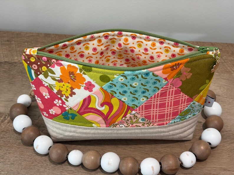 K&ouml;nnte beinhalten: Ein Rei&szlig;verschlusst&auml;schchen mit einer Patchwork-Au&szlig;enseite aus verschiedenen Blumen-, Karomustern und geometrischen Designs in Orange, Pink, Gelb, Blau und Gr&uuml;n. Das Innenfutter hat einen kleinen Blumendruck und der Boden besteht aus beigem Stoff.
