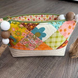 K&ouml;nnte beinhalten: Ein Rei&szlig;verschlusst&auml;schchen mit einem Patchwork-Au&szlig;enmaterial aus bunten Stoffquadraten mit Blumen-, Paisley- und Karomustern. Der Boden besteht aus gestepptem cremefarbenem Stoff. Zwei Jute-Quasten mit wei&szlig;en und braunen Holzperlen sind an den Zippern befestigt.