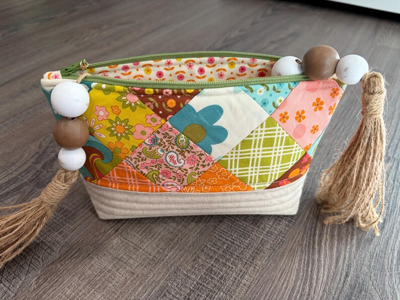 K&ouml;nnte beinhalten: Ein Rei&szlig;verschlusst&auml;schchen mit einem Patchwork-Au&szlig;enmaterial aus bunten Stoffquadraten mit Blumen-, Paisley- und Karomustern. Der Boden besteht aus gestepptem cremefarbenem Stoff. Zwei Jute-Quasten mit wei&szlig;en und braunen Holzperlen sind an den Zippern befestigt.