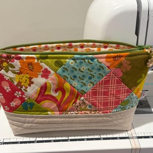 K&ouml;nnte beinhalten: Ein Rei&szlig;verschlusst&auml;schchen mit einem Patchwork-Design aus verschiedenen floralen und geometrischen Stoffquadraten in Orange, Pink, Blau und Gr&uuml;n. Der untere Teil besteht aus beigem Stoff mit horizontalen Steppn&auml;hten. Ein kleiner Blumenanh&auml;nger.