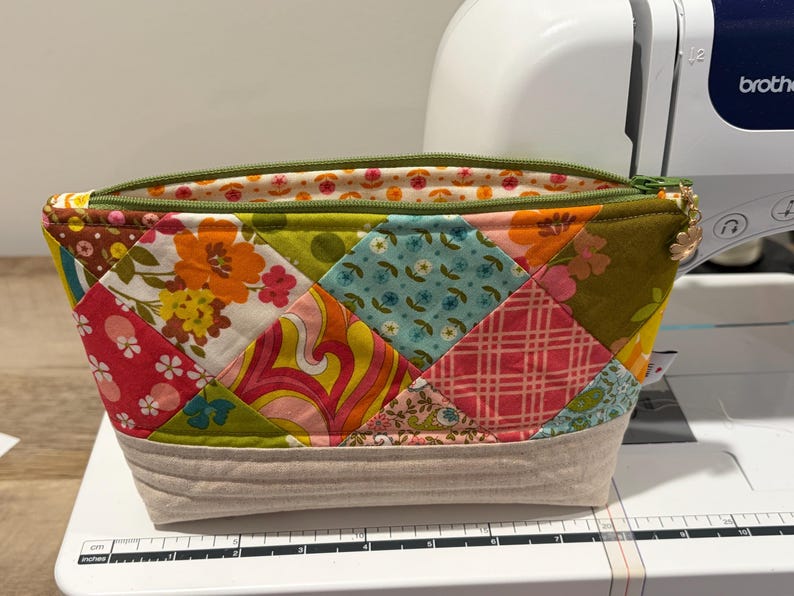 K&ouml;nnte beinhalten: Ein Rei&szlig;verschlusst&auml;schchen mit einem Patchwork-Design aus verschiedenen floralen und geometrischen Stoffquadraten in Orange, Pink, Blau und Gr&uuml;n. Der untere Teil besteht aus beigem Stoff mit horizontalen Steppn&auml;hten. Ein kleiner Blumenanh&auml;nger.