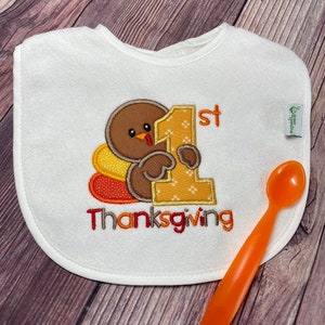 Puede incluir: Un babero blanco con un pavo marr&oacute;n bordado que sostiene un gran n&uacute;mero uno amarillo, con el texto 1st Thanksgiving debajo. Una peque&ntilde;a etiqueta de green sprouts est&aacute; en el lateral. Una cuchara de pl&aacute;stico naranja est&aacute; colocada al lado.