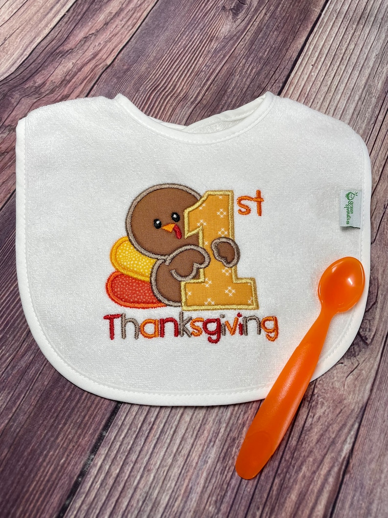 Puede incluir: Un babero blanco con un pavo marr&oacute;n bordado que sostiene un gran n&uacute;mero uno amarillo, con el texto 1st Thanksgiving debajo. Una peque&ntilde;a etiqueta de green sprouts est&aacute; en el lateral. Una cuchara de pl&aacute;stico naranja est&aacute; colocada al lado.
