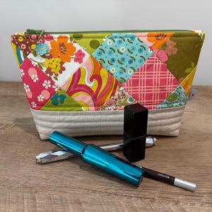 K&ouml;nnte beinhalten: Ein Rei&szlig;verschlusst&auml;schchen mit Patchwork-Design aus Blumen- und geometrischen Mustern in Orange, Pink, Gr&uuml;n und Blau sowie einem hellgrauen Boden. Ein kleiner blauer Blumenanh&auml;nger h&auml;ngt am Rei&szlig;verschluss. Dazu geh&ouml;ren eine t&uuml;rkisfarbene Mascara, ein schwarzer Lippenstift und ein silberner Eyeliner.