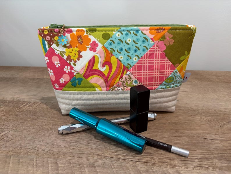 K&ouml;nnte beinhalten: Ein Rei&szlig;verschlusst&auml;schchen mit Patchwork-Design aus Blumen- und geometrischen Mustern in Orange, Pink, Gr&uuml;n und Blau sowie einem hellgrauen Boden. Ein kleiner blauer Blumenanh&auml;nger h&auml;ngt am Rei&szlig;verschluss. Dazu geh&ouml;ren eine t&uuml;rkisfarbene Mascara, ein schwarzer Lippenstift und ein silberner Eyeliner.