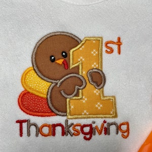 Puede incluir: Un babero blanco con un bordado de un pavo marr&oacute;n que sostiene un gran n&uacute;mero 1 amarillo con el texto 1st Thanksgiving. El pavo tiene plumas de cola con patrones amarillos y naranjas. El bordado utiliza hilo marr&oacute;n, amarillo, naranja y rojo.