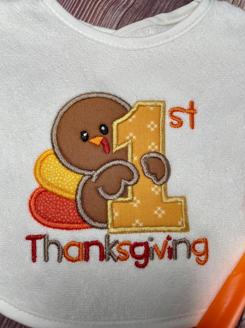 Puede incluir: Un babero blanco con un bordado de un pavo marr&oacute;n que sostiene un gran n&uacute;mero 1 amarillo con el texto 1st Thanksgiving. El pavo tiene plumas de cola con patrones amarillos y naranjas. El bordado utiliza hilo marr&oacute;n, amarillo, naranja y rojo.