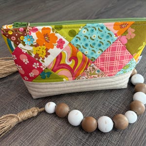 May include: A zippered pouch featuring a patchwork design of colorful floral and geometric fabric squares in shades of orange, pink, blue, and green. The base is quilted beige fabric, and a blue flower charm hangs from the green zipper pull.