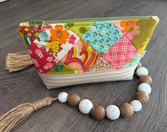 May include: A zippered pouch featuring a patchwork design of colorful floral and geometric fabric squares in shades of orange, pink, blue, and green. The base is quilted beige fabric, and a blue flower charm hangs from the green zipper pull.