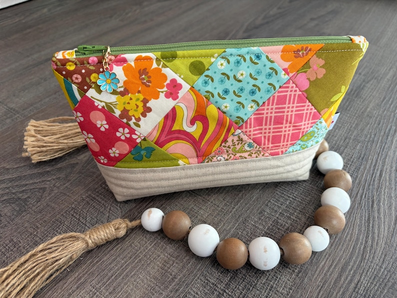K&ouml;nnte beinhalten: Eine Rei&szlig;verschlusstasche mit einem Patchwork-Design aus bunten floralen und geometrischen Stoffquadraten in Orange, Pink, Blau und Gr&uuml;n. Die Basis besteht aus gestepptem beigem Stoff und ein blauer Blumenanh&auml;nger h&auml;ngt am gr&uuml;nen Rei&szlig;verschluss.